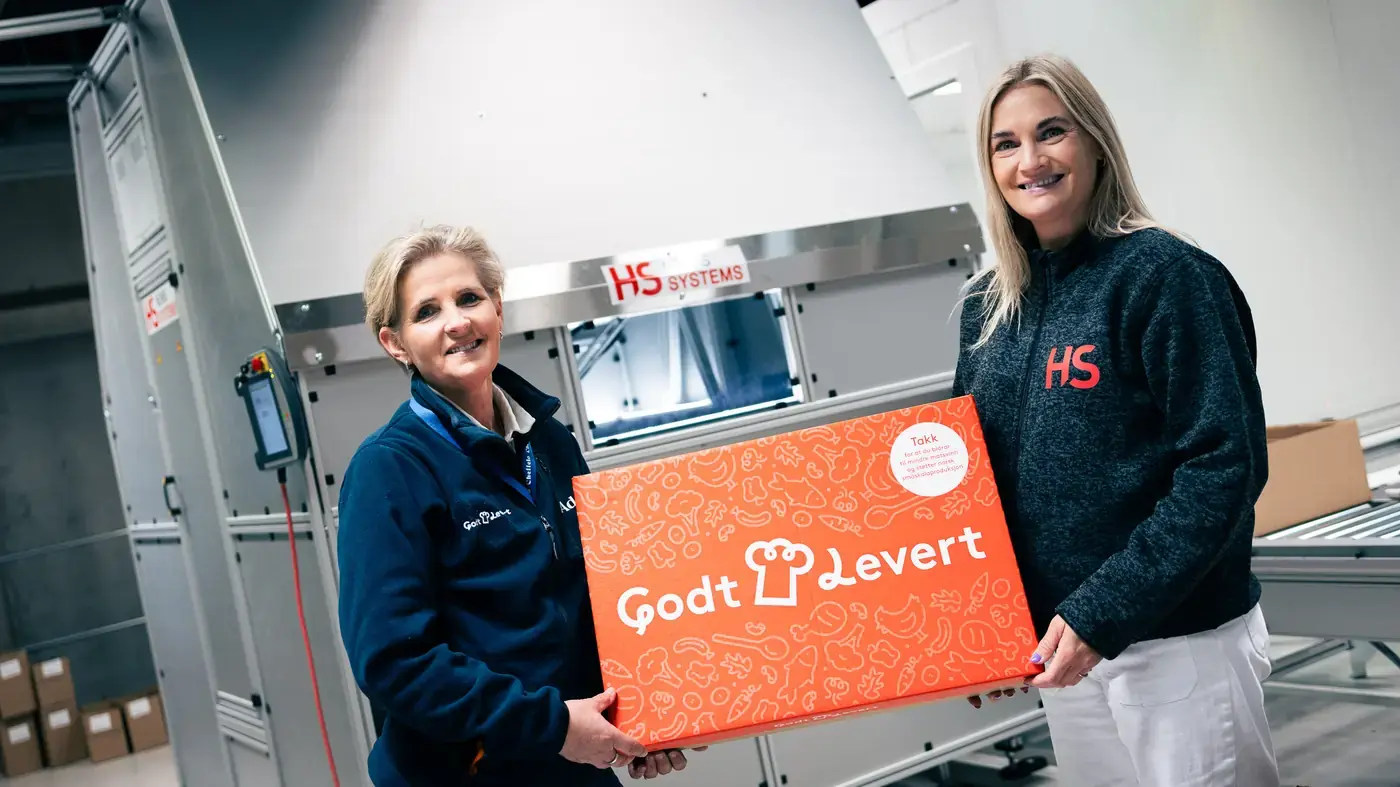 Two people holding a Godt Levert meal-kit box in front of the HS pick robot